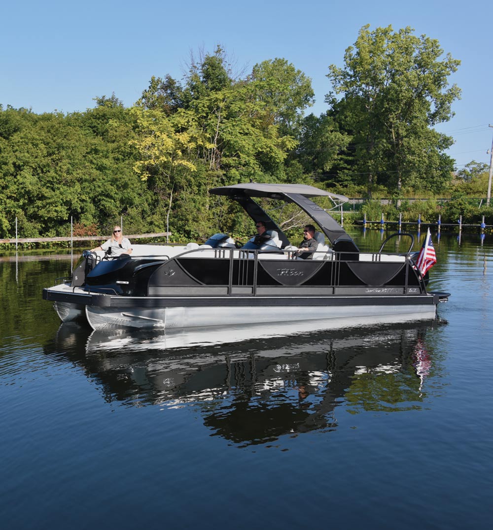 A black pontoon boat with three people on a calm lake, surrounded by trees, and an American flag at the back.