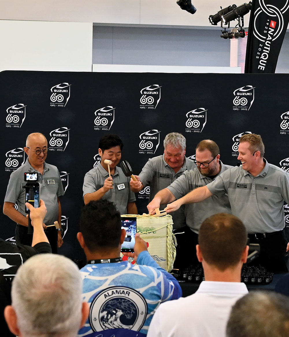 Five men in gray shirts conduct a ceremonial sake cask opening in front of a "Suzuki 60 1965-2025" backdrop.