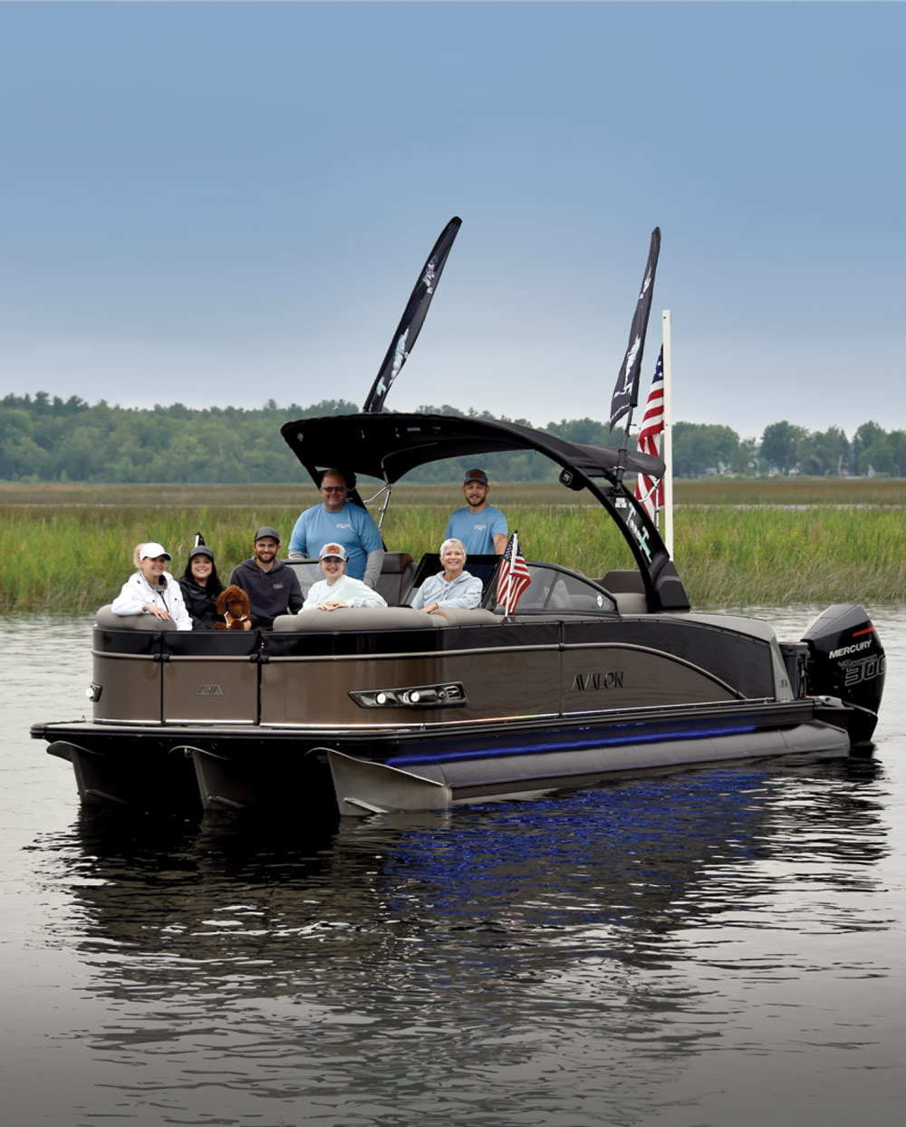 A pontoon boat with seven people on calm water, surrounded by greenery, under a cloudy sky.