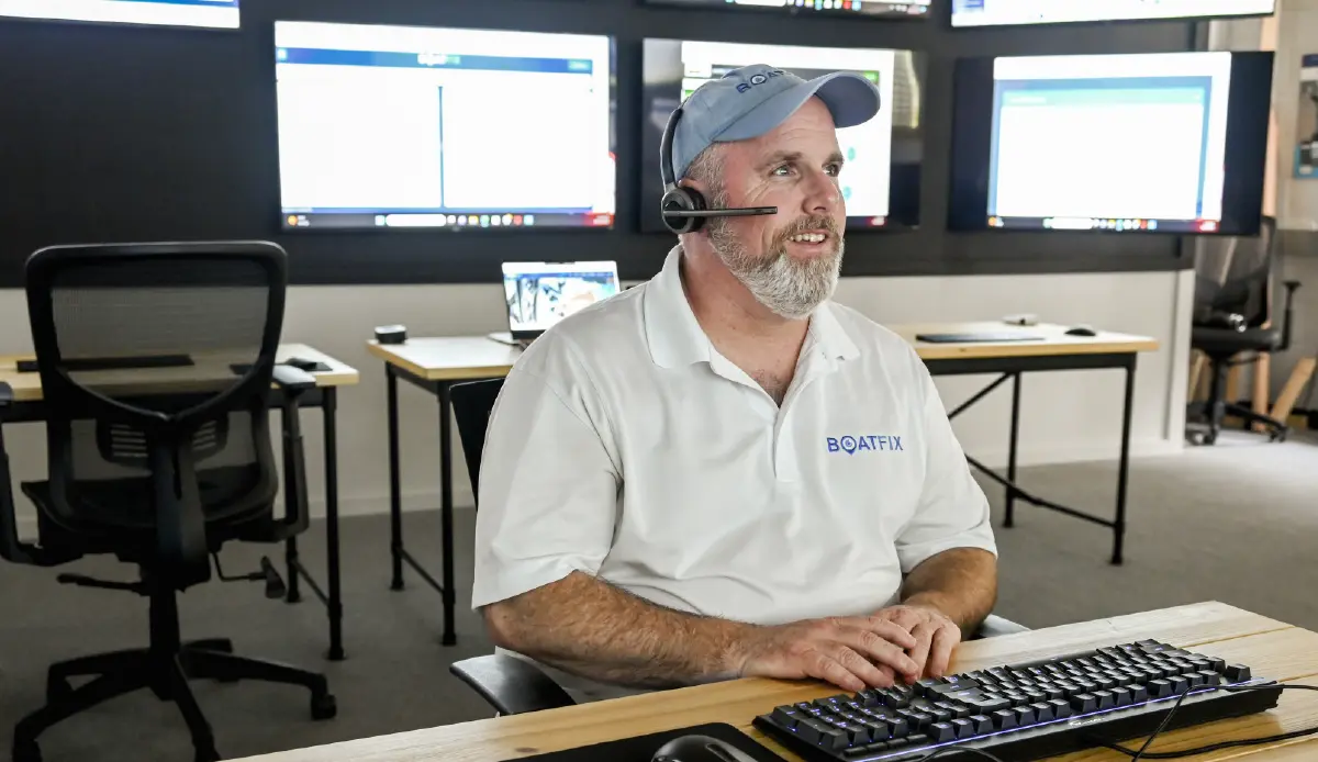 Landscape orientation photograph of a bearded man with a headset and Boat Fix company polo top t-shirt sitting at a call center desk smiling with a computer keyboard in front of him as well as a computer mouse nearby as he looking towards multiple computer screens in the foreground and behind him in the background are other additional desks, computer chairs, and computer screens
