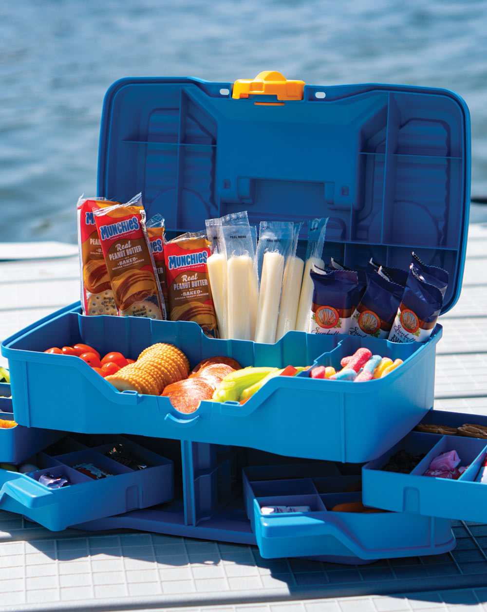 Open blue storage box filled with various snacks, placed on a deck by the water.