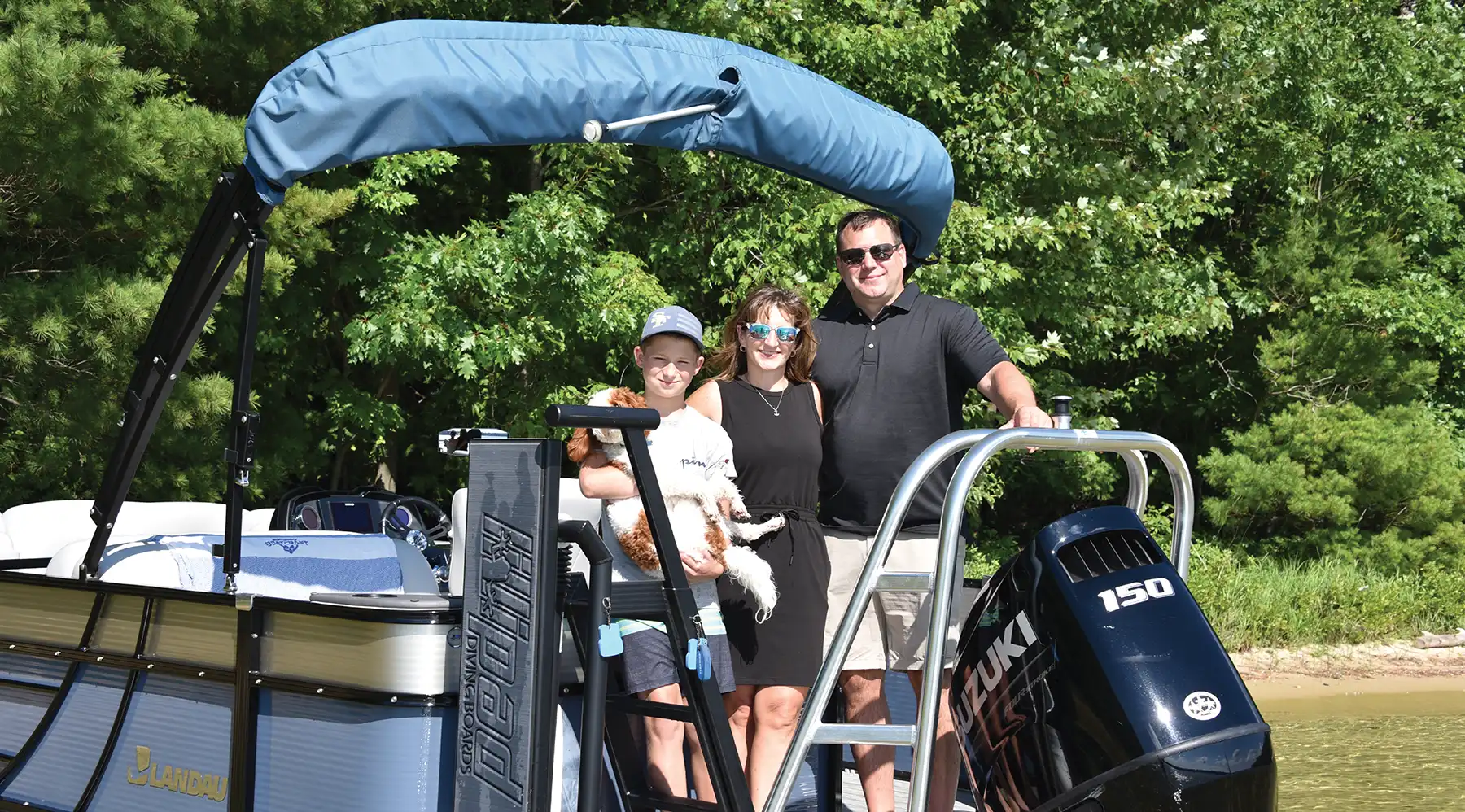 A family and their dog standing on a pontoon boat with a blue canopy and a Suzuki 150 motor, near a wooded shoreline.
