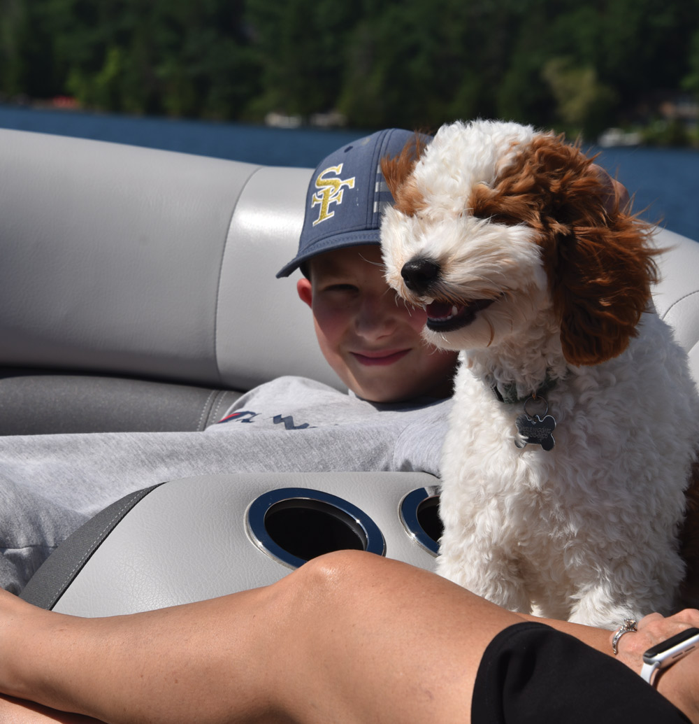 A boy wearing an "SF" cap sits with a small curly-haired dog on a boat, with a lake and trees in the background.