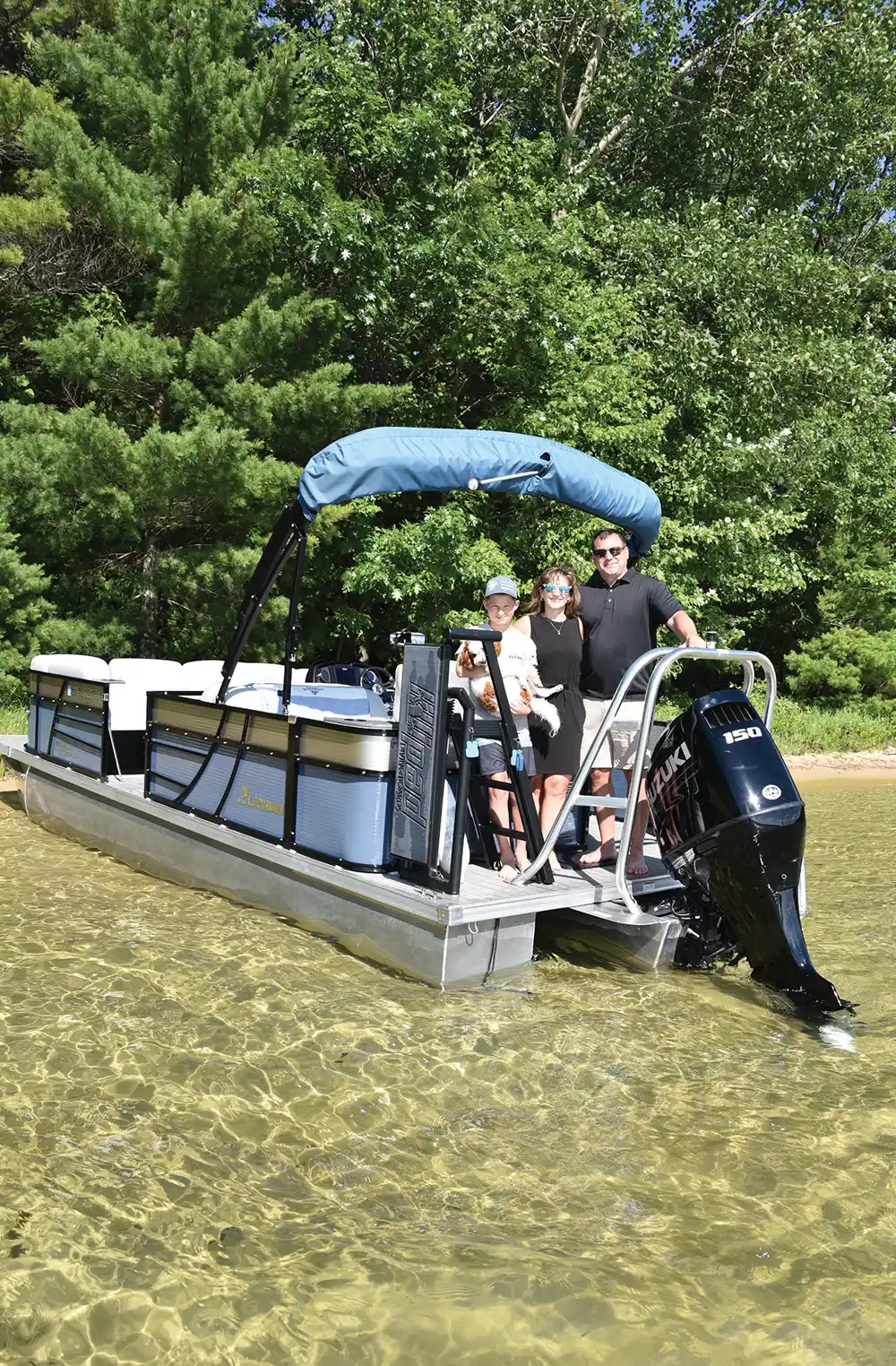 Three people on a pontoon boat near a sandy shoreline with trees in the background.