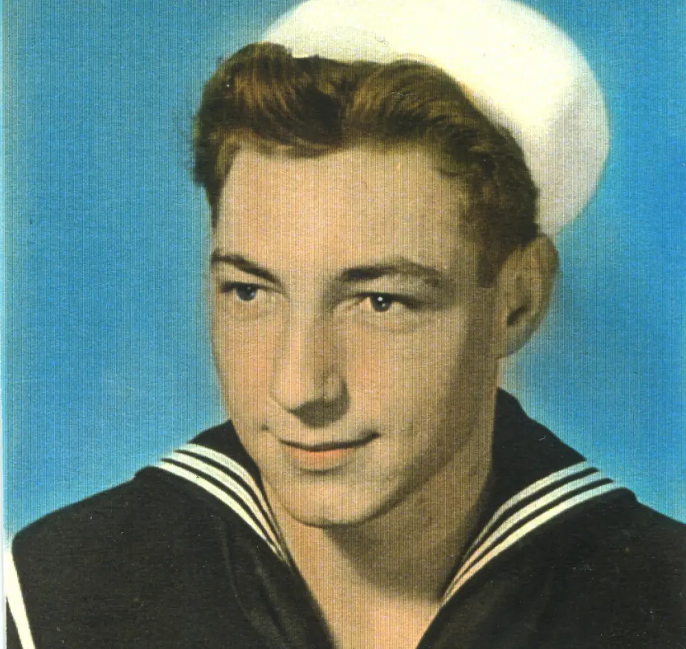 Retro vintage old school portrait orientation headshot photograph of a younger Ronald Fisher in his Navy sailor uniform outfit attire featuring either a presumed black or dark navy blue top that contains white stripes and his white sailor hat hanging on top of his dark brown hair