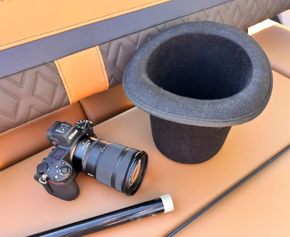 Portrait orientation outdoor photograph close-up view of a black Nikon camera with a large lens resting on a brown colored pontoon motorboat vehicle couch seat as this camera is next to a bottom upside down facing orientation black top hat; There happens to also be a partial visible black stick pole containing a white closed cap on it nearby the camera and top hat as well as orange, black, dark chocolate brown, and dark charcoal grey textured upholstery which is part of the pontoon motorboat vehicle couch seat material