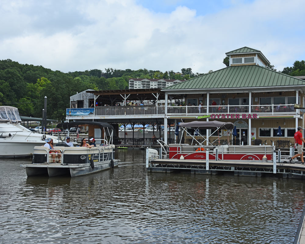 Boats at a marina with a two-story restaurant and forested hills in the background.