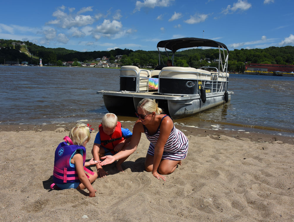 Children and an adult playing on a sandy beach with a pontoon boat in the background.