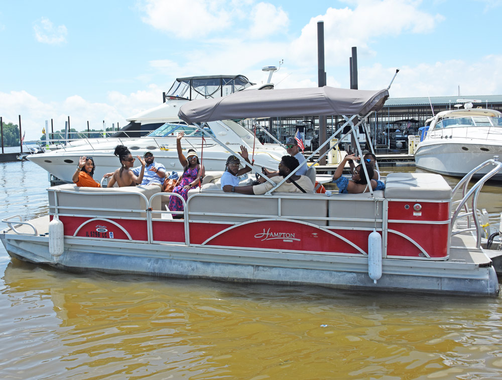 A pontoon boat with people waving is docked at a marina, surrounded by larger yachts.