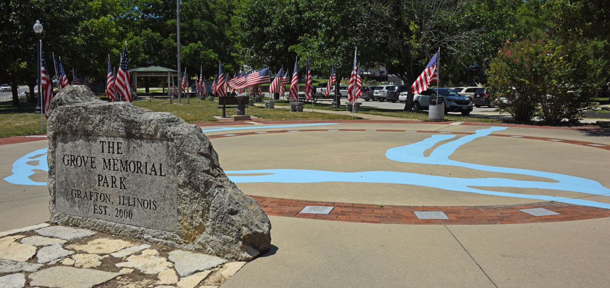 A memorial park with a large engraved rock, American flags, and a painted pavement design.