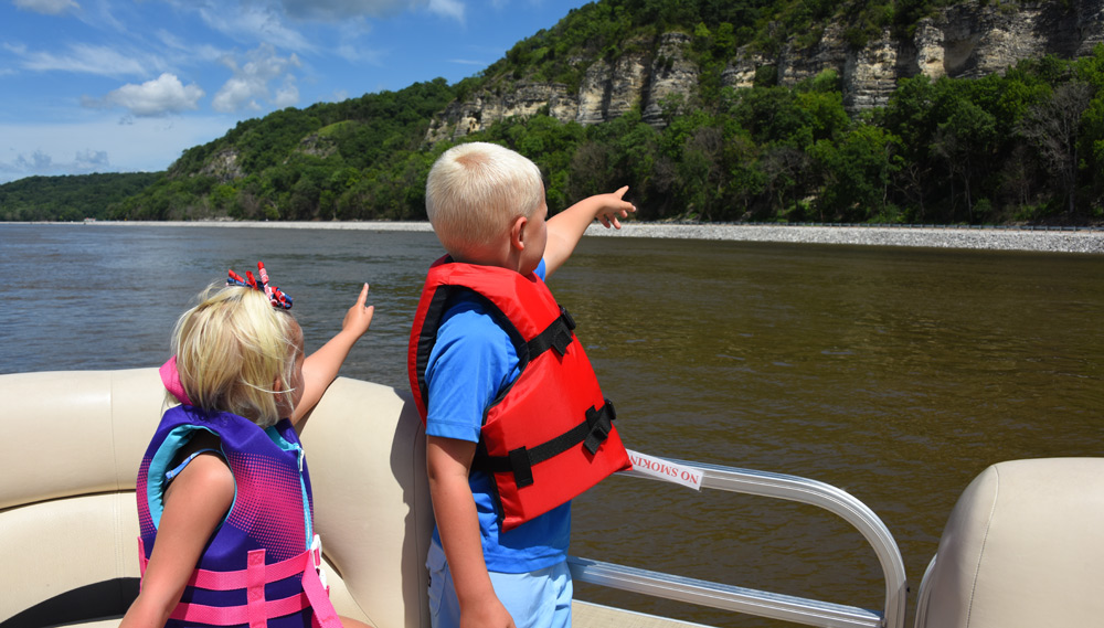 Two children in life jackets point toward a riverbank with cliffs and trees.