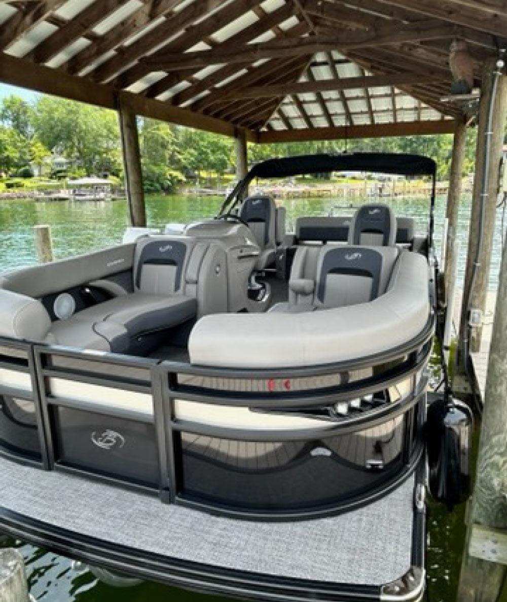 A dark grey pontoon boat with black accents, featuring grey and black seating, is docked under a wooden boathouse.