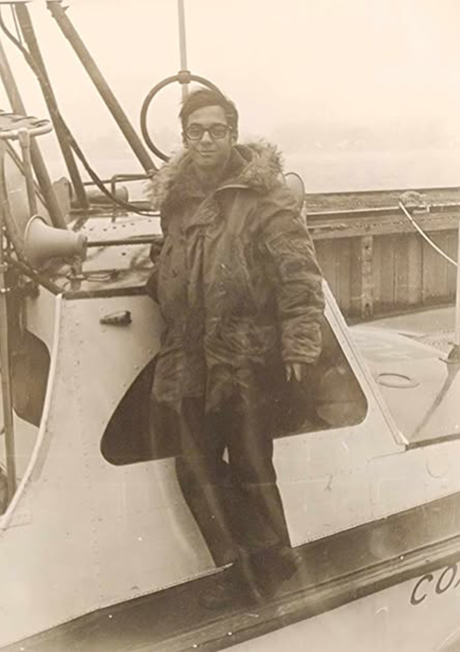 A vintage, sepia-toned photo of a young man wearing glasses and a heavy, fur-lined winter coat, standing on the deck of a boat.