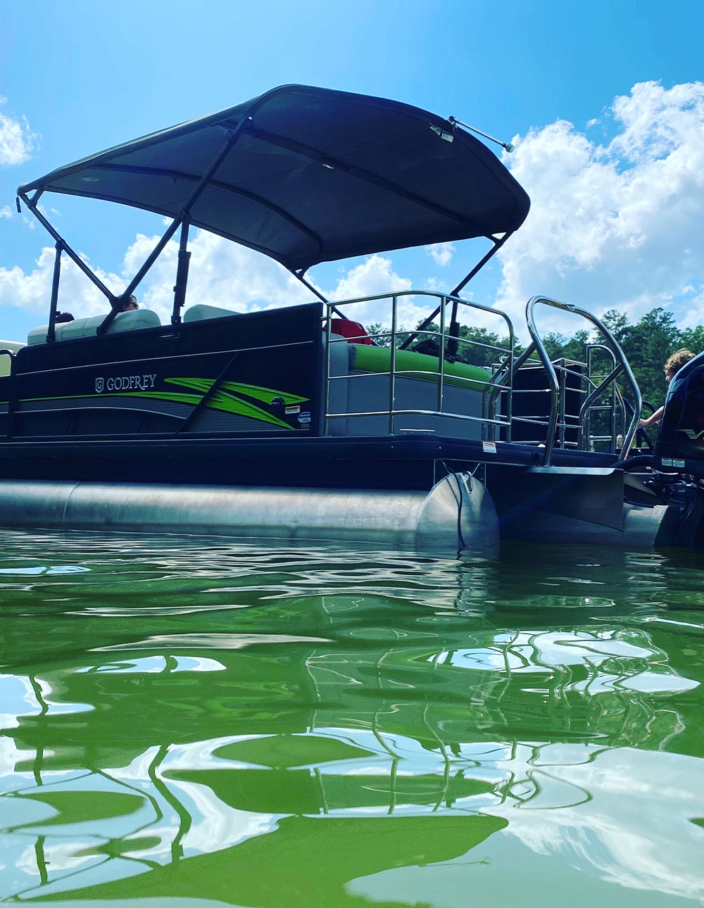 A low-angle view from the water of a dark-colored pontoon boat with green accents and a canopy, floating on greenish water under a blue sky with clouds.