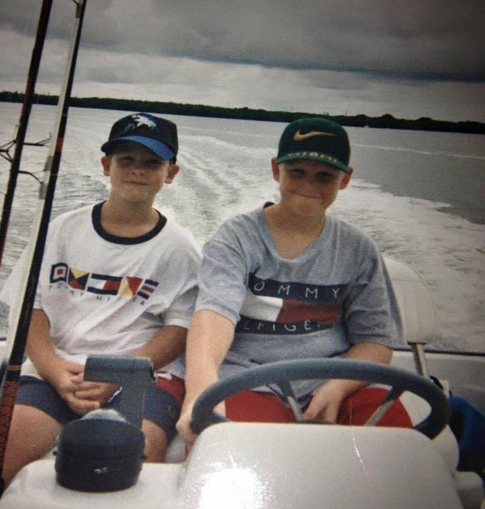 two young boys, both wearing caps, smile while sharing the seat at the helm of a pontoon in sail