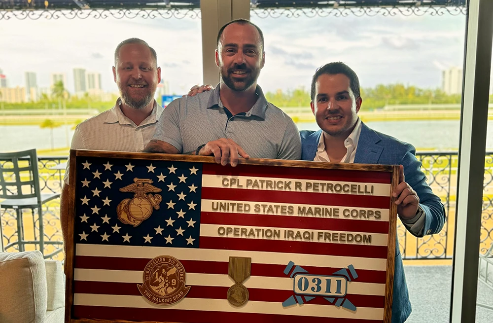 Three men are holding and presenting a large, wooden plaque designed like an American flag. The plaque is dedicated to "CPL PATRICK R PETROCELLI" and commemorates his service in the "UNITED STATES MARINE CORPS" during "OPERATION IRAQI FREEDOM," and features various military insignia.