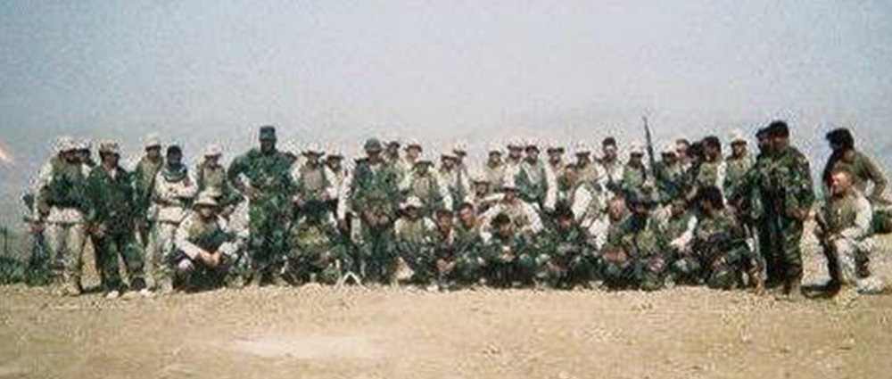 A grainy, wide shot of a large group of soldiers in camouflage uniforms and combat gear posing for a photo in a desert environment.