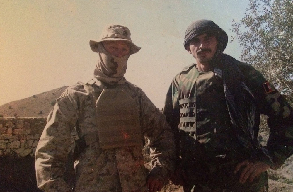 A soldier wearing a boonie hat, face covering, and full camouflage gear stands next to another soldier in a different uniform and a head wrap. They are posing together outdoors in a dry, hilly environment.