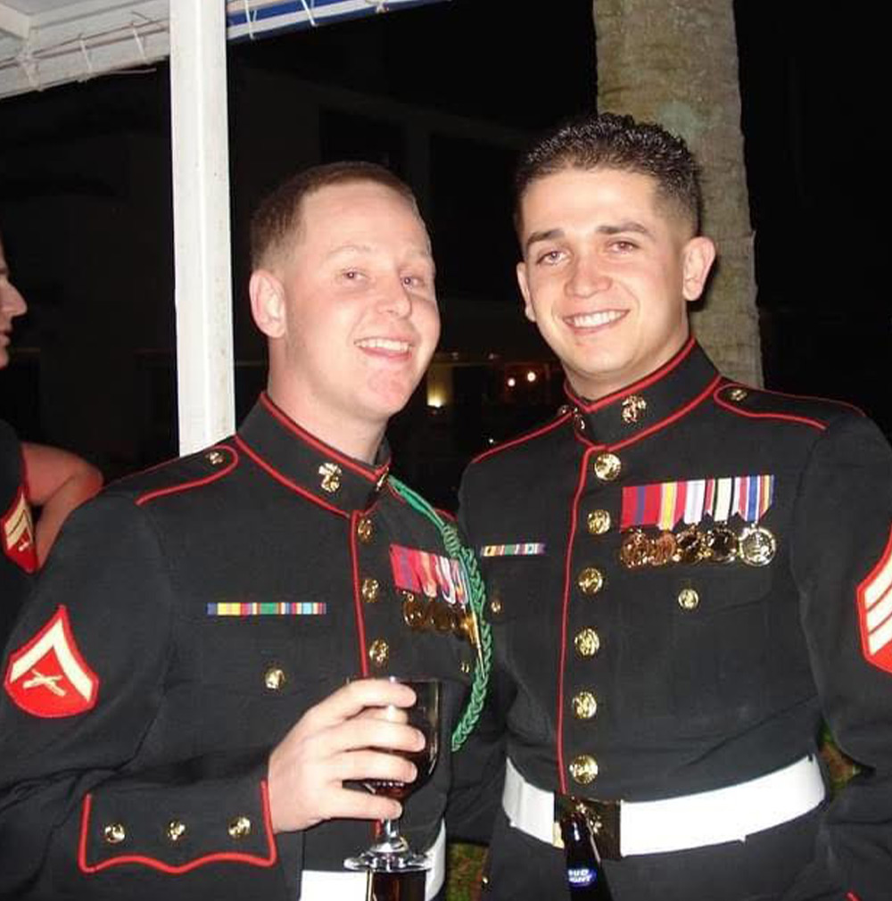 Two men in United States Marine Corps Dress Blue uniforms are smiling for a photo at a nighttime event. The Marine on the left holds a glass of wine, and both men are decorated with ribbons and medals.