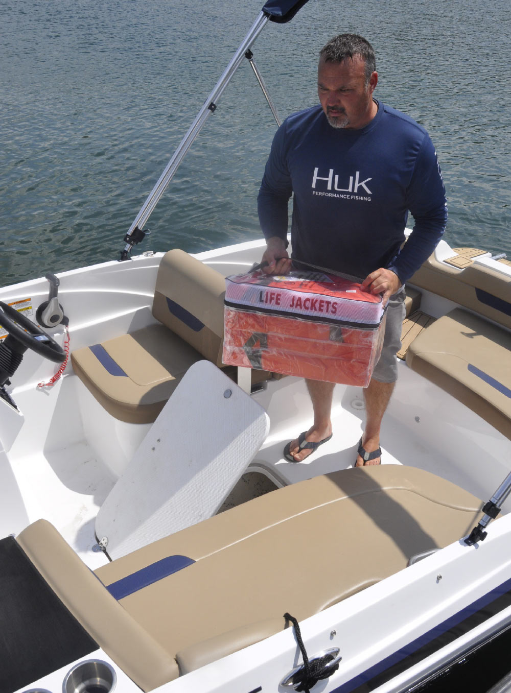 A man stands on a boat holding a transparent bag filled with orange life jackets.