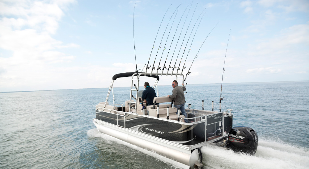 A black and white pontoon boat, labeled "ANGLER QUEST" and "MERCURY 150," speeds across calm water under a partly cloudy sky. Numerous fishing rods are mounted on the boat, with several extended upwards from a rack. Two men are on board: one is seated at the helm, and the other stands near the stern.