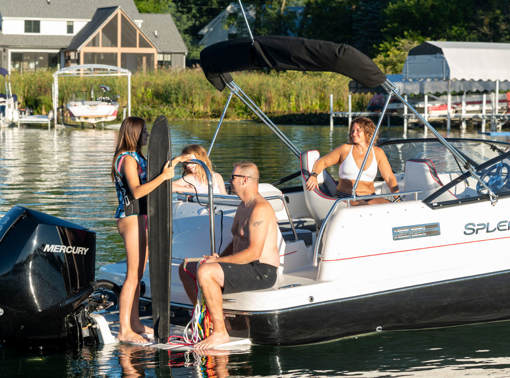 Four people on a motorboat at a lake. A girl holds a water ski, while others sit on the boat.