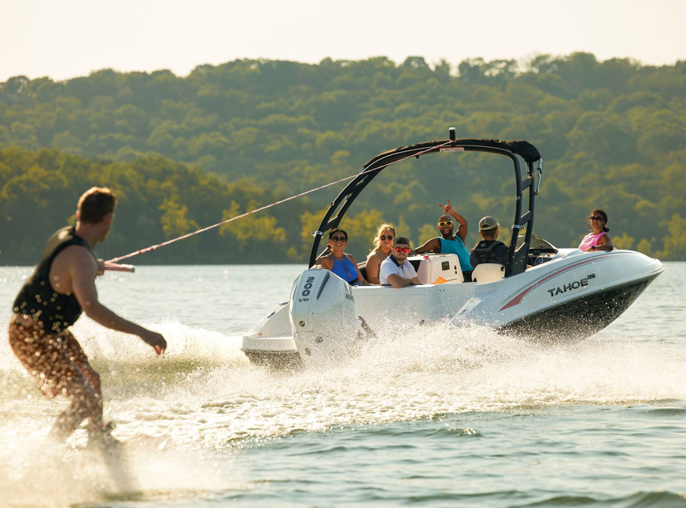 A speedboat on a lake towing a wakeboarder, with trees in the background.