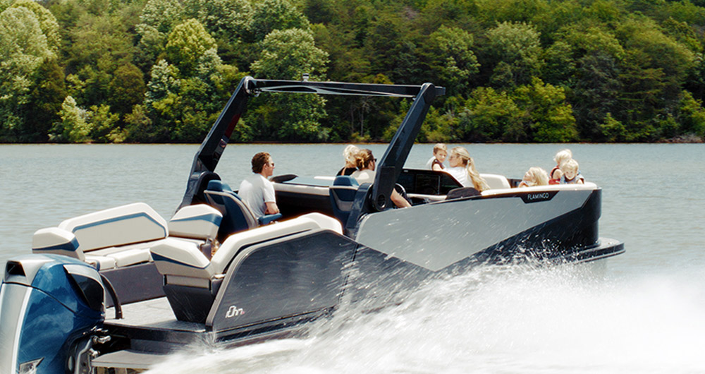 A rOhm pontoon full of people is speeding across a lake, with trees in the background.