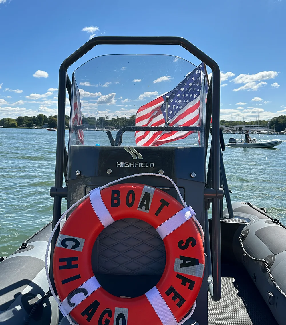 A life preserver with "BOAT SAFE CHICAGO" written on it is attached to the front of a boat, with an American flag flying behind it and water and other boats in the background.