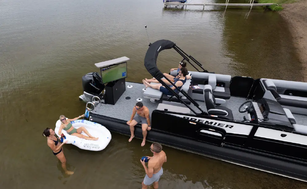 An aerial outdoor landscape orientation view of the 250 Solaris RS (Rear Social) pontoon motor boat from Premier Pontoons with a black and gray colored design floating near a sandy shore on a lake; Several people are on board and others are in the shallow water, some relaxing on a white inflatable float; The boat has a TV screen visible at the back, and a remote dock is situated in the far distance