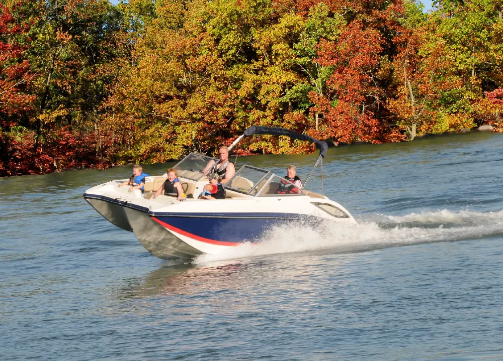 Outdoor portrait orientation photograph of a white, red, and blue colored pontoon motorboat with a canopy which happens to be moving across a lake, creating a wake behind it; A man is steering the boat, and three children are seated in the front while there happens to be another adult passenger behind the driver as well as another adult passenger seated in front of the driver's viewpoint; The shore in the background is lined with trees in fall colors, including beautiful color shades of reds, oranges, and yellows
