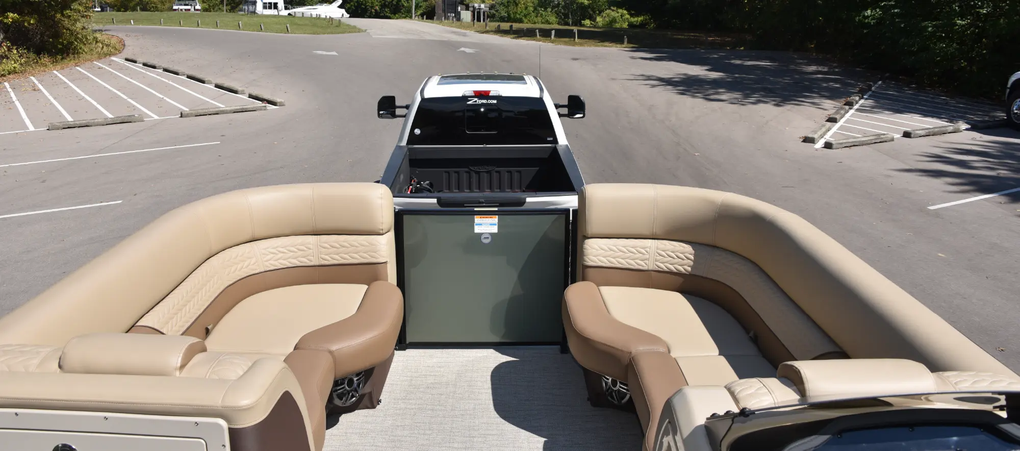 A view from the back of a pontoon motorboat, showing its light brown or tan beige colored cushioned seating; In the center of the boat, there is a small doorway path gate entrance; A white pickup truck is visible ahead, attached to the boat's trailer; The truck is backed into a boat launch parking lot, with trees and a few other vehicles in the background