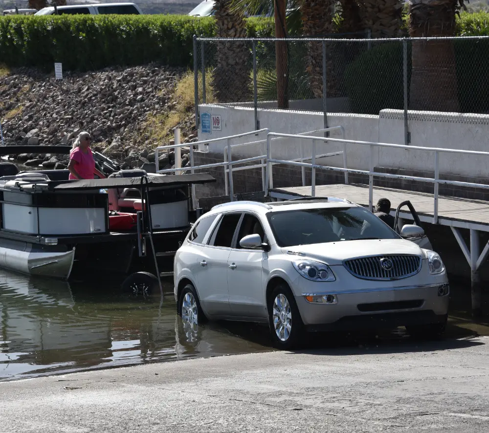 A white SUV is backed into a lake on a concrete boat ramp, preparing to launch a dark grey, black, and chrome colored pontoon motorboat; A woman in a pink shirt is on the boat and another person is visible by the driver's car side door of the white SUV; A dock and chain link fence are in the background, with the shoreline visible on the left