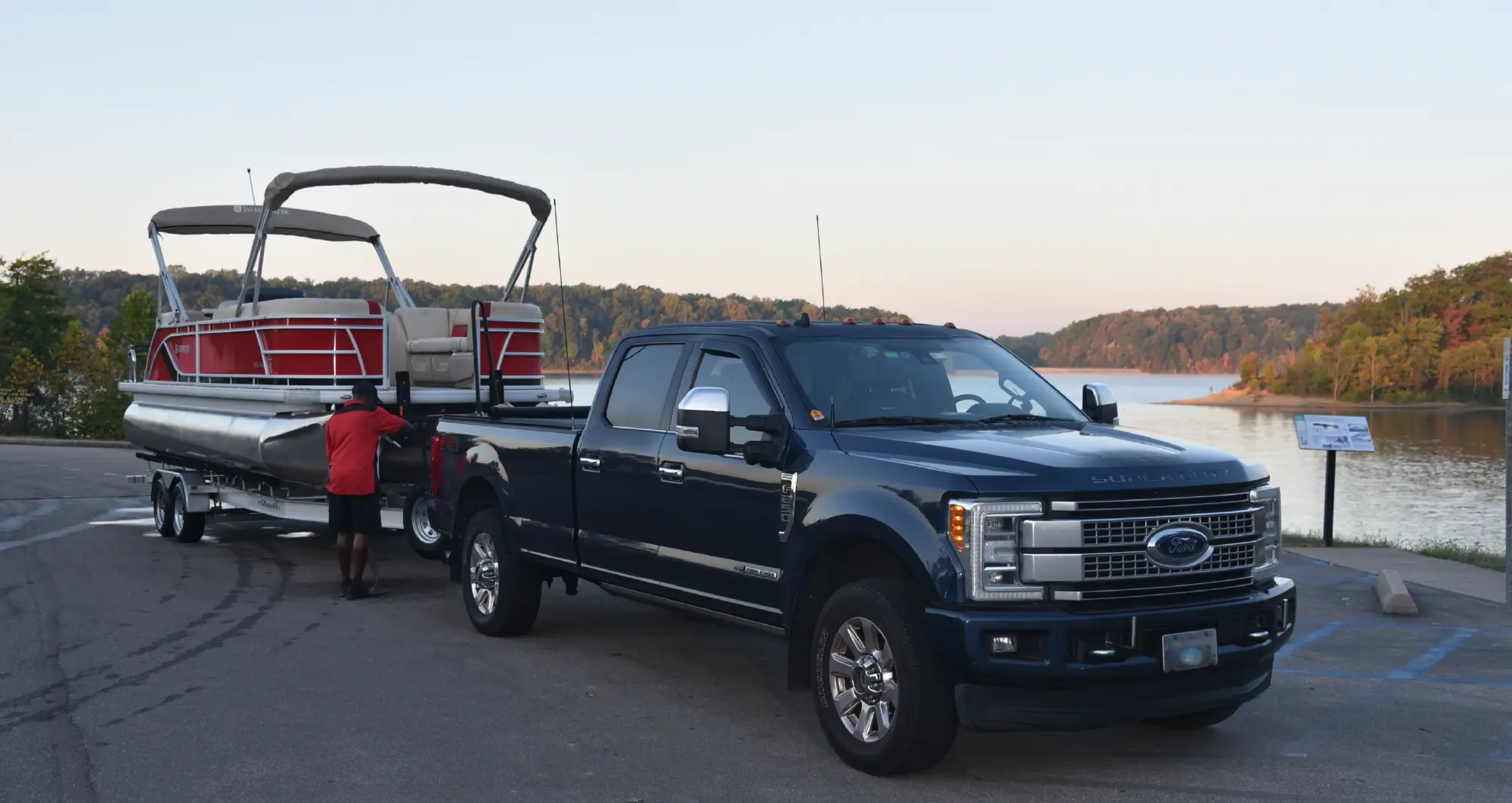 A dark navy blue Ford Super Duty pickup truck is backed up to a boat launch ramp, with a red/beige colored pontoon motorboat on a trailer attached to it; A person in a red polo top shirt is standing near the back of the boat; The boat is facing a lake surrounded by trees under a bright, early evening sky