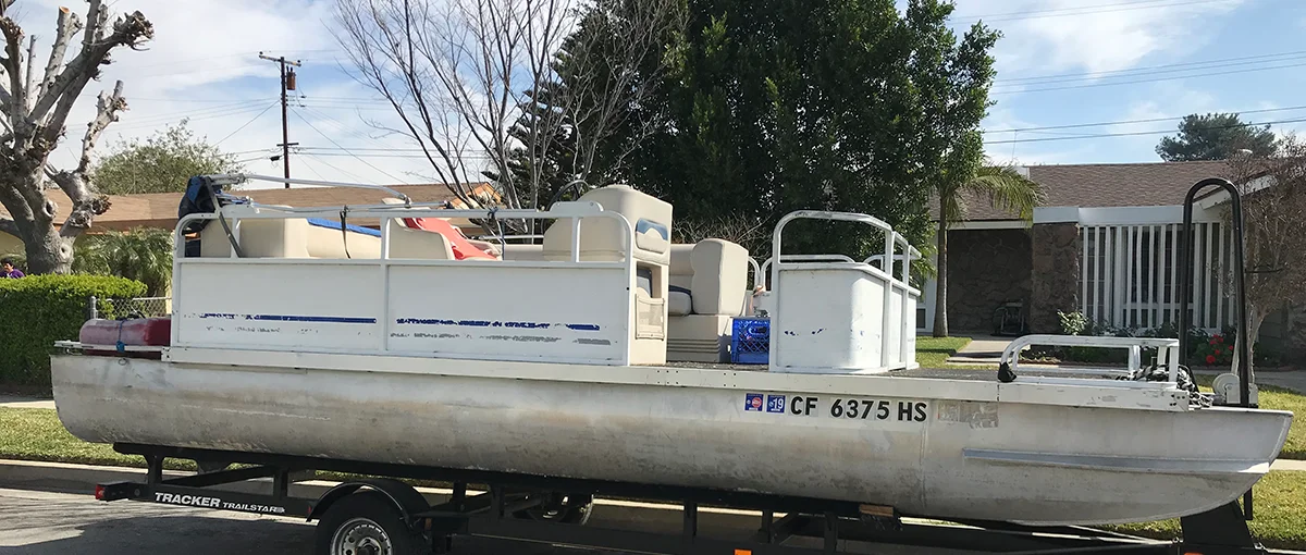 A pontoon boat in poor condition, showing peeling paint and discoloration, sitting on a Tracker Trailstar trailer.