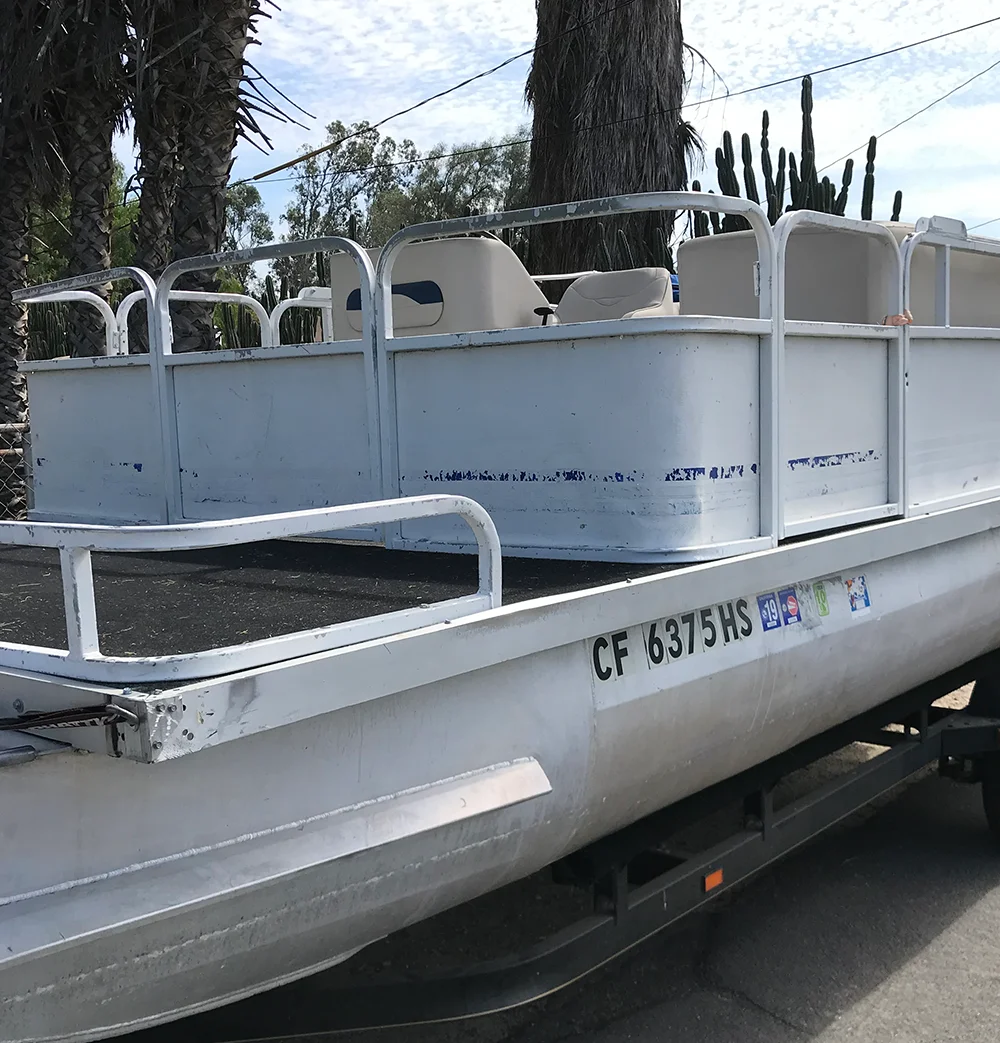 A close-up side view of a pontoon boat on a trailer, showing its worn-out white paint, old seating, and aluminum hull.