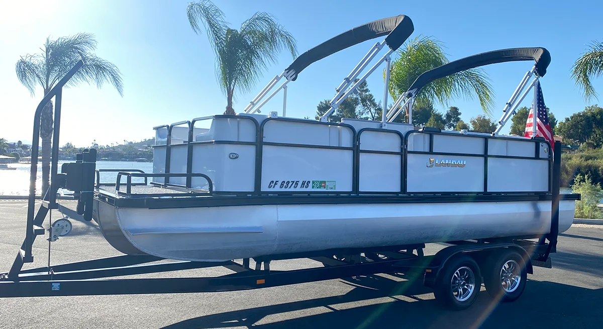 A newly restored pontoon boat with a dual black canopy, black railings, and a small American flag sits on a trailer in front of a lake.