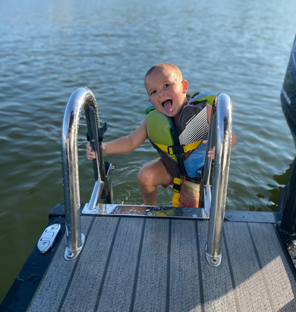 A joyful young boy in a life jacket climbs a ladder onto a pontoon boat, sticking his tongue out and smiling at the camera.