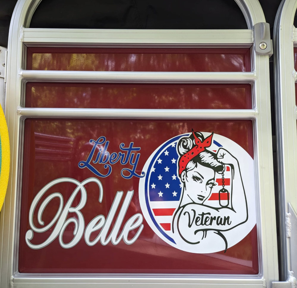 A close-up photograph of two decals. The one on the left says "Liberty Belle" in a flowing white script with a blue outline. The decal on the right is a circular sticker featuring a woman's face and arm, reminiscent of the "Rosie the Riveter" icon. 
