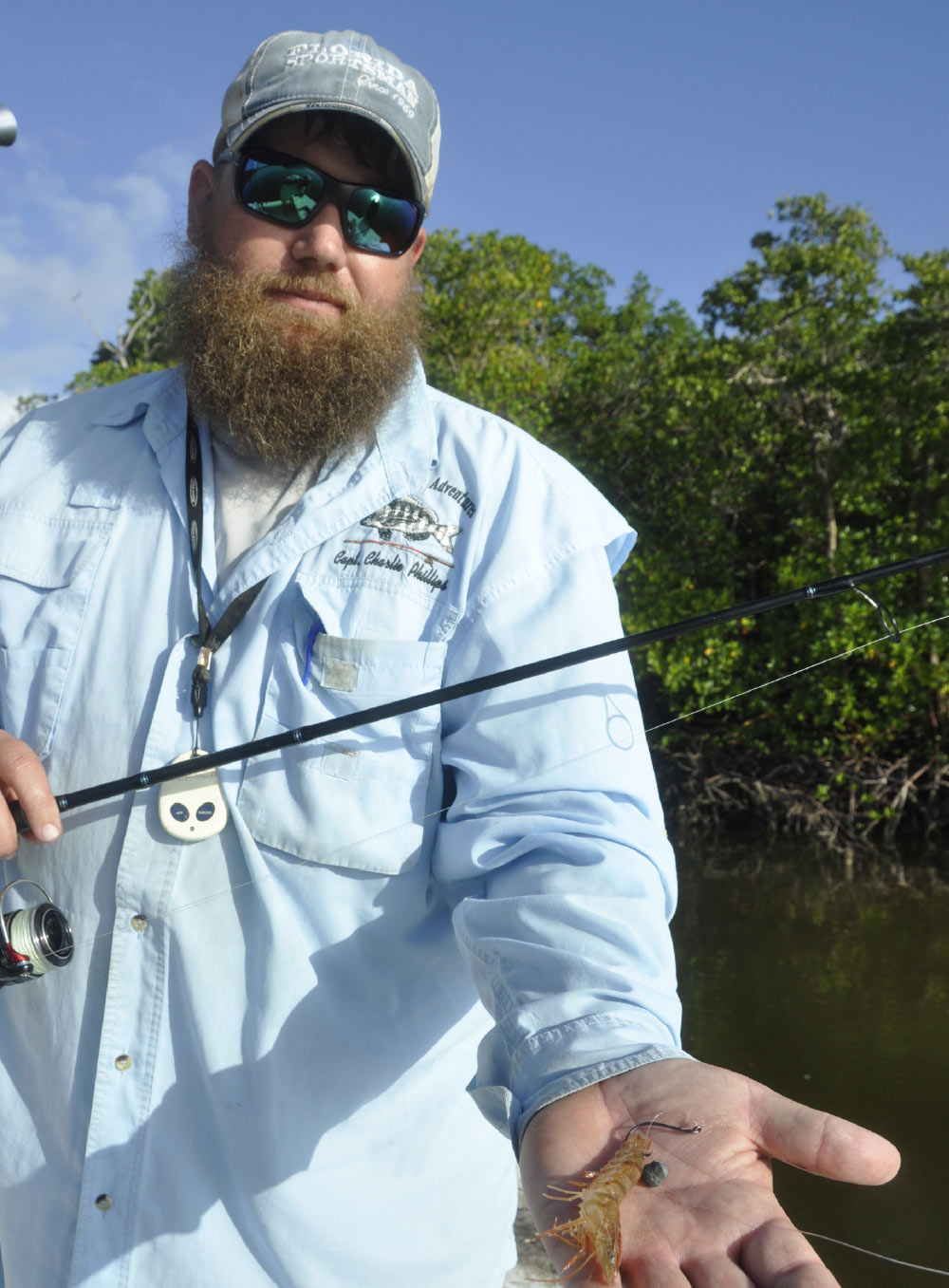 A low-angle, vertical photograph of a bearded man with mirrored sunglasses and a baseball cap holding a fishing rod and a small shrimp in his open palm. 