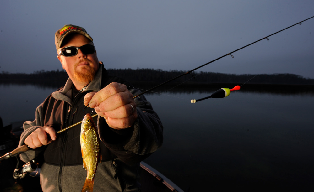 A wide-angle, horizontal photograph of a man on a boat, holding a fishing rod with a small fish on the hook. 