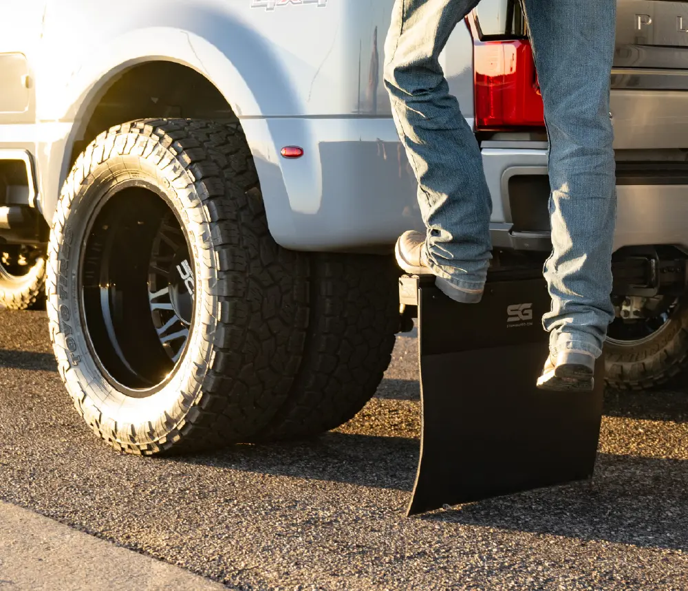 A close-up shot of the back wheel and a black Step N Guard modular STEP/mudflap SYSTEM of a gray colored pickup truck; The mudflap has a logo on it; A person's legs in blue jeans and shoes are visible as they step up onto the truck's rear bumper; The scene is outdoors on a paved road, and the low sun casts a warm light