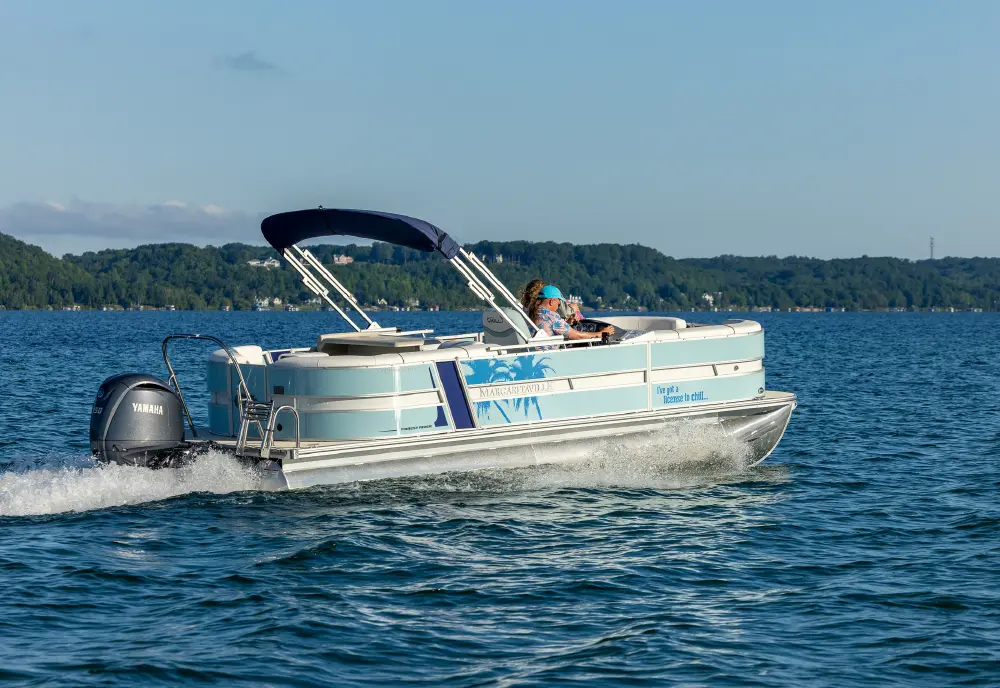 A light sky blue colored pontoon motorboat with a dark navy blue canopy is moving across a large lake; A person is sitting in the driver's seat; A Yamaha engine is visible at the back and the side of the boat has a palm tree graphic and a MARGARITAVILLE logo plus a quote that says I've got a reason to chill!; The shoreline, covered with trees, is visible in the background under a blue sky