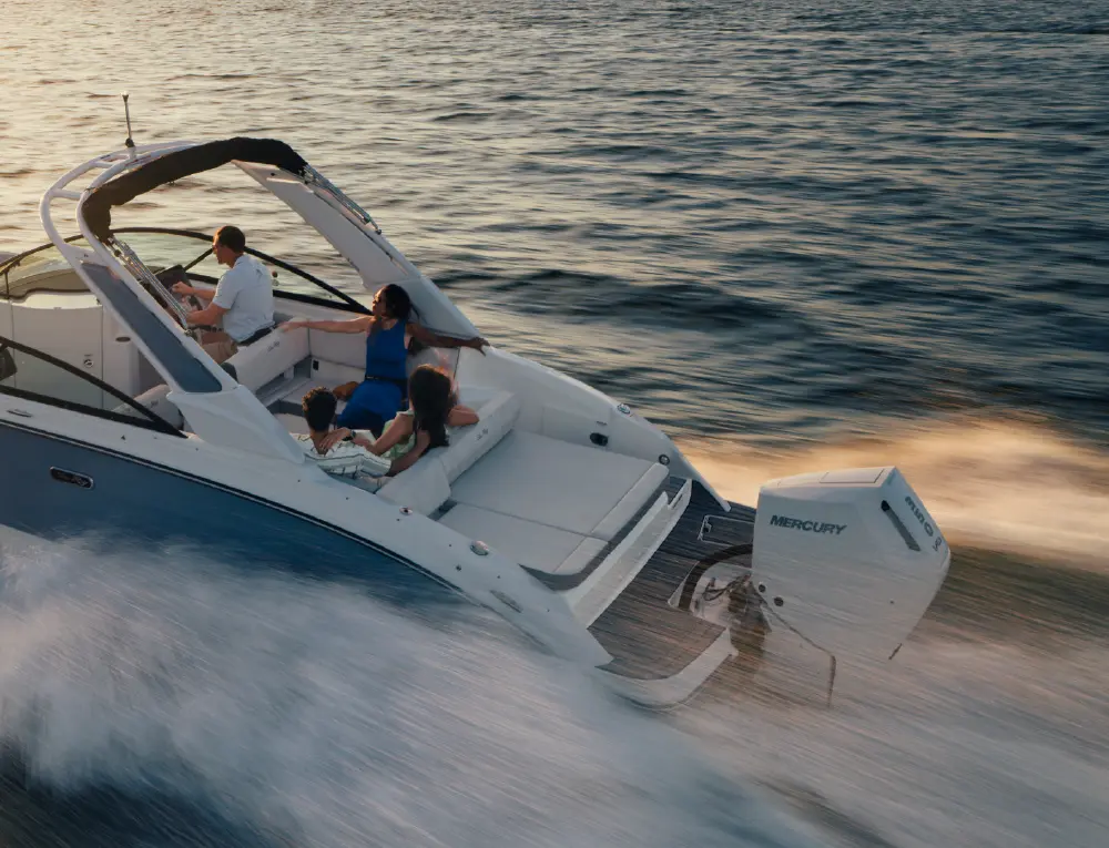 An aerial photo from behind a white and dark baby blue colored pontoon motorboat moving across a body of water, creating a wake; A family of four is on board: a man is steering from the driver's seat, a woman is sitting on the passenger side, and two individuals are seated in the back; A white Mercury engine is visible at the stern; The water is calm with a golden sunset light
