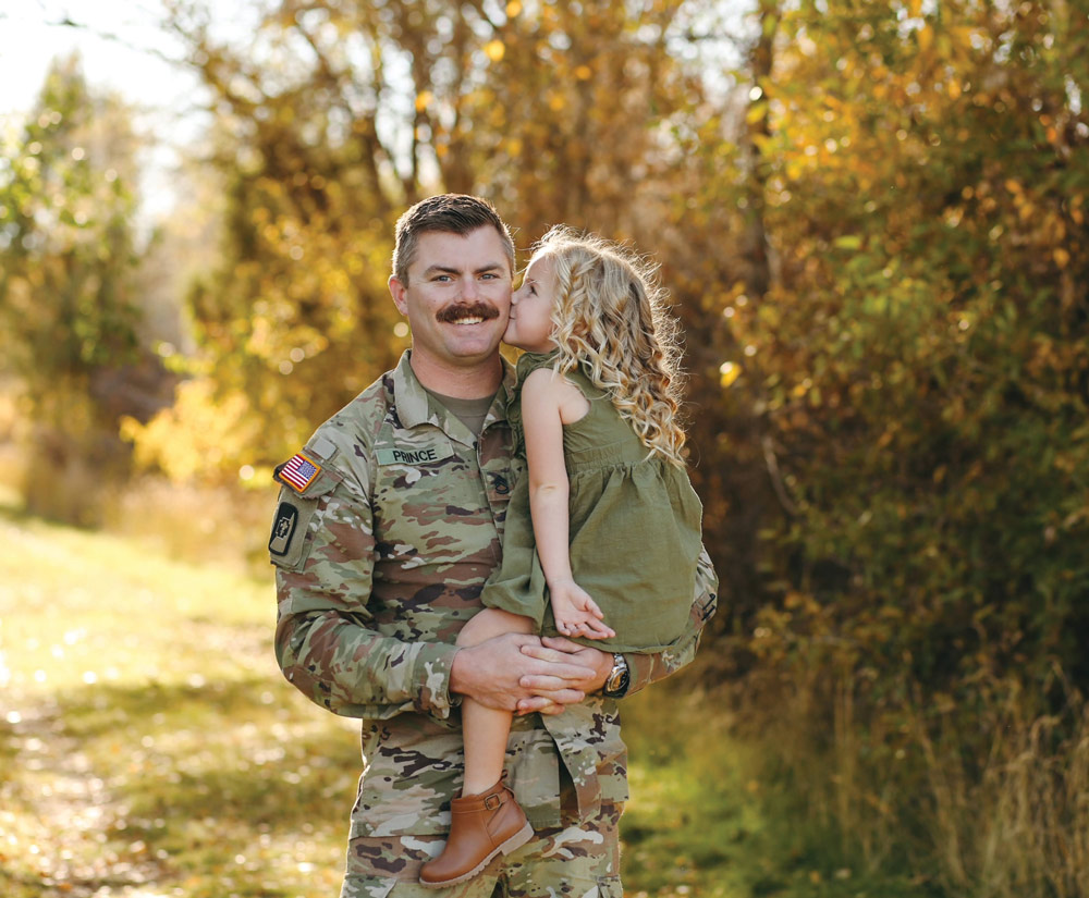 A U.S. Army soldier with a mustache is holding a young girl with blonde curly hair in a green dress and brown boots. The girl is kissing the soldier on his cheek as they both smile at the camera. They are standing outside in a park setting with trees. The soldier's name tape says PRINCE.