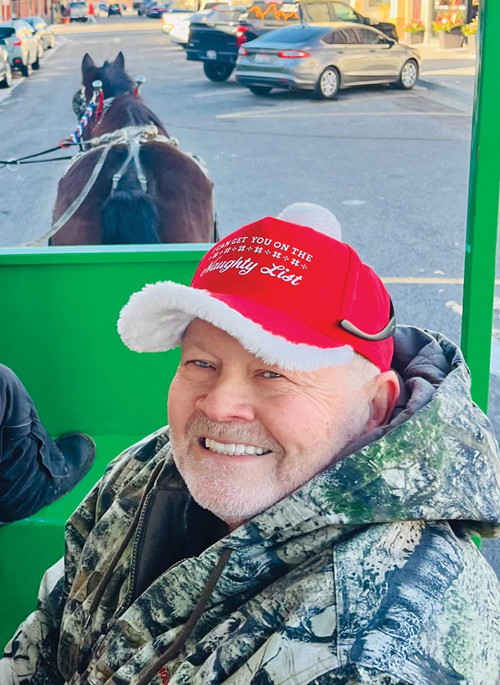 A close-up of a smiling older man with a white beard, wearing a camouflage hoodie and a red Santa hat with a white pom-pom. The hat has text on it that says, "I'd Get You On The Naughty List." He is sitting in a horse-drawn carriage.