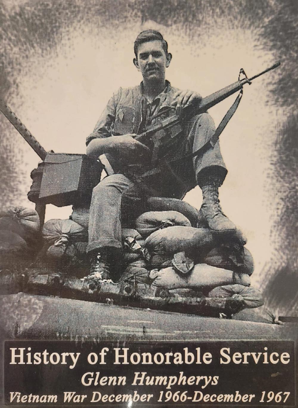 A black and white photo of Glenn Humpherys, a Vietnam War veteran, sitting on a pile of sandbags with his rifle. He's wearing a military uniform, boots, and is looking directly at the camera. Below the image, text reads: "History of Honorable Service," "Glenn Humpherys," and "Vietnam War December 1966-December 1967."