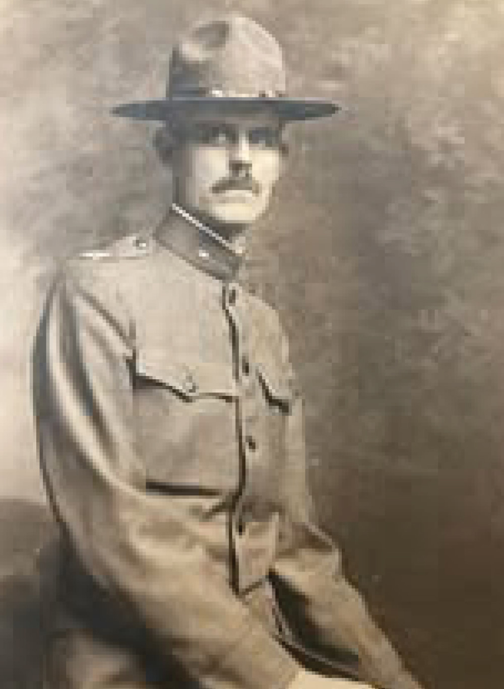 A sepia-toned vintage portrait of a man in a military uniform from the early 20th century. He has a mustache and is wearing a wide-brimmed hat. He is looking at the camera with a serious expression.