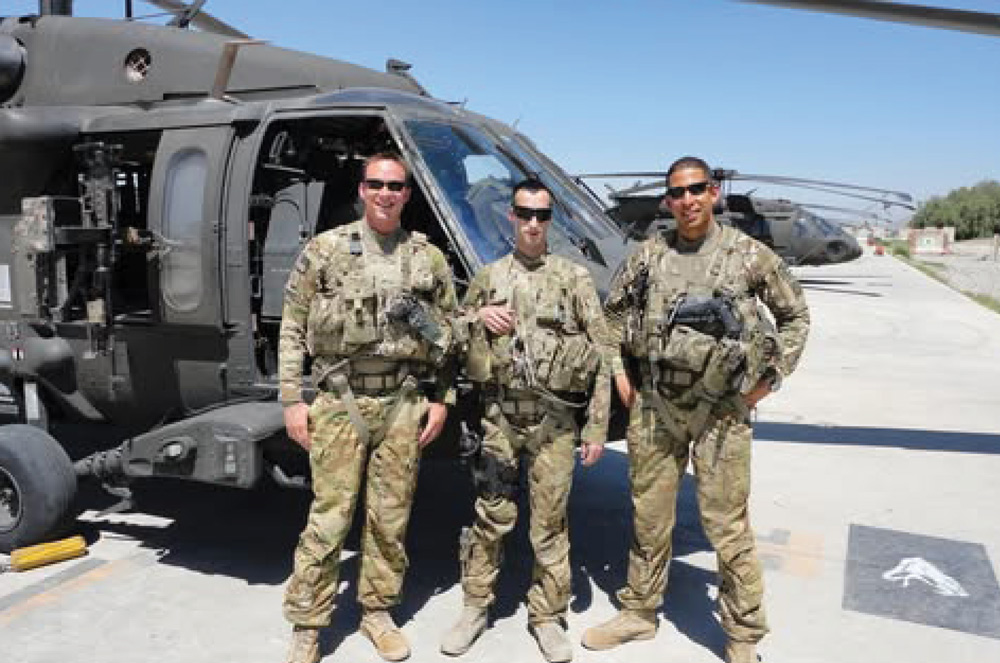 Three U.S. Army soldiers in camouflage uniforms and sunglasses are standing side-by-side in front of a Black Hawk helicopter. The soldiers are wearing tactical vests and are posing for the camera on a sunny day.