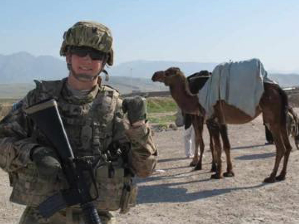 A photo of a U.S. soldier in full combat gear, including a helmet and sunglasses, holding a rifle and giving a slight smile to the camera. Behind him, a camel with a pack is standing in a desert-like, mountainous landscape.